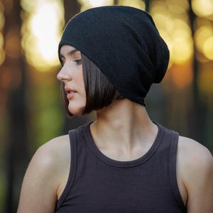 May include: A black beanie hat and a dark gray tank top. The beanie is made of a soft material and is worn on the head. The tank top is sleeveless and has a rounded neckline. The background is blurred with warm tones.