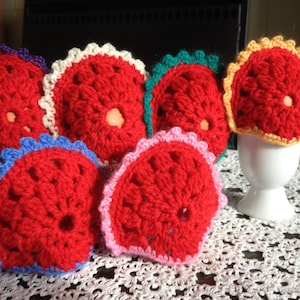 May include: Six crocheted egg cozies in various colors, including red, white, yellow, green, and blue. The cozies are shaped like a flower with a hole in the center for the egg. They are sitting on a white crocheted doily.