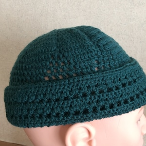 May include: A dark green crocheted beanie hat with a textured pattern. The hat is worn on a mannequin head.