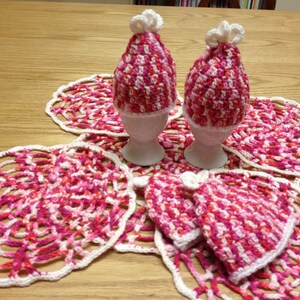 May include: Two pink and white crocheted egg cozies on a white crocheted doily.