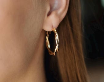 May include: Close-up of a gold hoop earring with a twisted design. The earring is a classic accessory, featuring a polished finish and a secure clasp. The jewellery is shown against a blurred background.