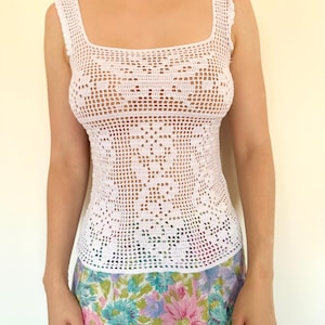 Pode incluir: Um top de crochê branco com decote quadrado e um padrão detalhado em renda. O top tem alças com babados e uma silhueta ajustada. A modelo usa uma saia floral com flores rosa, azul e roxa.
