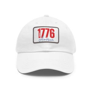Puede incluir: Una gorra de béisbol blanca con un parche rectangular en la parte delantera. El parche tiene un borde gris oscuro y muestra el texto rojo 1776 sobre la palabra America escrita en letra cursiva azul. La gorra tiene una visera curva y estructurada.