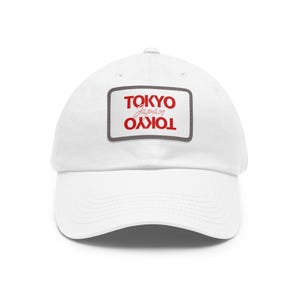 May include: A white baseball cap featuring a rectangular patch on the front. The patch has a dark gray border and displays the word TOKYO in red capital letters at the top and bottom, with the word Japan written in a red script font in the center.