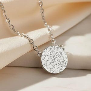 Puede incluir: Un collar plateado con un colgante circular. El colgante presenta un diseño abstracto detallado en un color más claro sobre un fondo más oscuro. La cadena es delicada y descansa sobre una tela blanquecina suave.