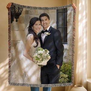 May include: A woven throw blanket featuring a wedding photo of a smiling couple. The bride wears a white gown and holds a bouquet. The groom is in a black suit. The blanket has a fringed edge.