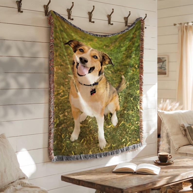 May include: Woven throw blanket featuring a smiling dog on a green grassy background. The dog is brown, tan, and white, wearing a black collar with a tag. The blanket is hanging on a white wall with decorative hooks.