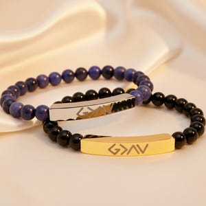 Könnte beinhalten: Zwei Armbänder mit Perlen. Ein Armband hat dunkelblaue Perlen und einen silbernen Balken mit einem geätzten Design. Das andere Armband hat schwarze Perlen und einen goldenen Balken mit den geätzten Buchstaben G>AV.