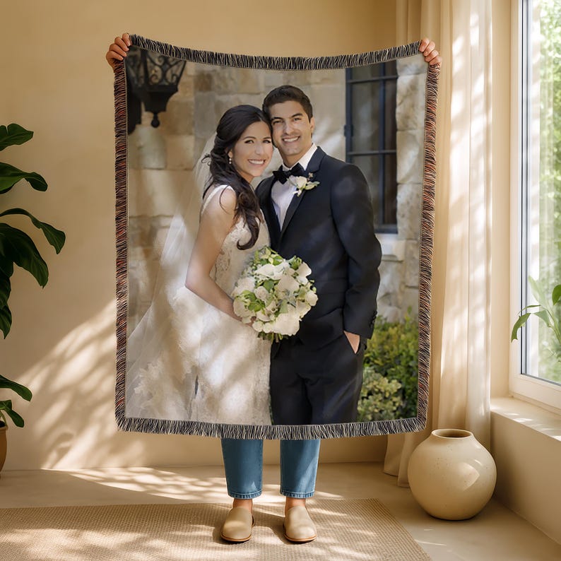 May include: A woven throw blanket featuring a wedding photo of a smiling couple. The bride wears a white gown and holds a bouquet. The groom is in a black suit. The blanket has a fringed edge.