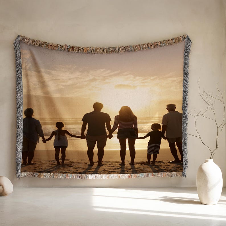 May include: Woven tapestry blanket featuring a family silhouetted against a sunset over the ocean. The image shows a family holding hands on a beach. The blanket has a fringed edge and is displayed on a wall.