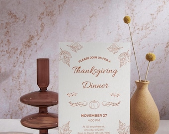 Puede incluir: Tarjeta de invitación a la cena de Acción de Gracias con ilustraciones de hojas de otoño y calabaza. La tarjeta es de color crema con texto marrón que dice "Please join us for a Thanksgiving Dinner" y detalles del evento. También hay un candelabro de cristal marrón y un jarrón con flores secas.