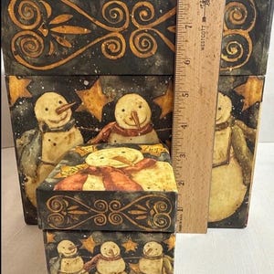 May include: Set of three decorative boxes featuring a winter snowman theme. The boxes are painted with a black background, gold stars, and whimsical snowmen wearing scarves. The boxes are different sizes, with the largest box measuring approximately 8 inches tall.