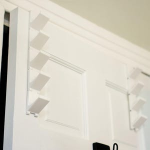 May include: White door with built-in, angled, white coat hooks. The hooks are mounted in a zig-zag pattern on the door. The door is part of a white door frame. A black object is visible at the bottom of the door.