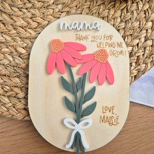 May include: Wooden plaque with the word "mama" in white script. The plaque features two pink flowers with orange centers and green stems. Text reads "Thank you for helping me grow!" and "Love, Maisie."
