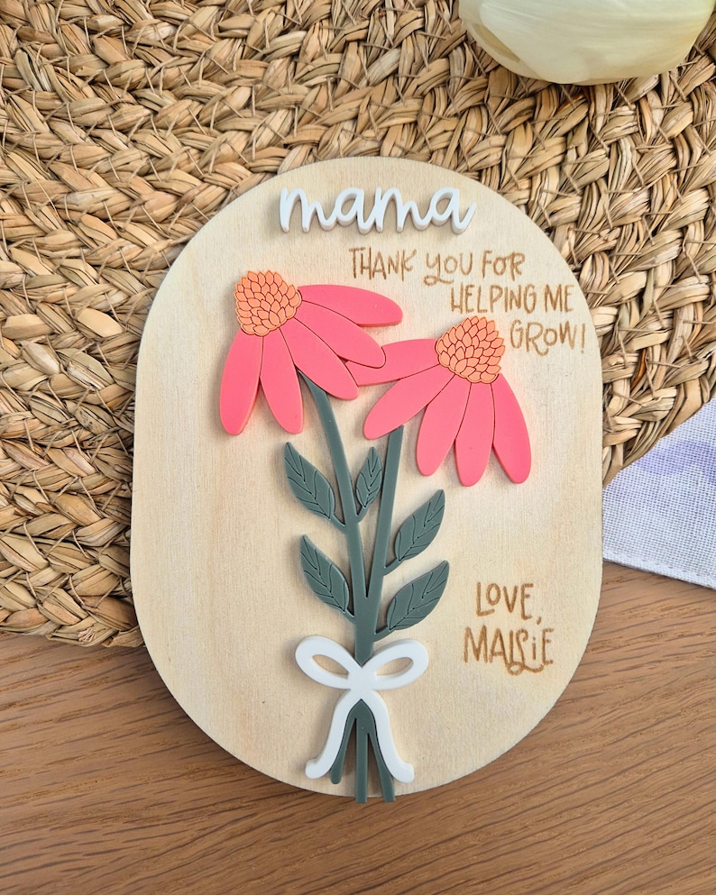 May include: Wooden plaque with the word "mama" in white script. The plaque features two pink flowers with orange centers and green stems. Text reads "Thank you for helping me grow!" and "Love, Maisie."