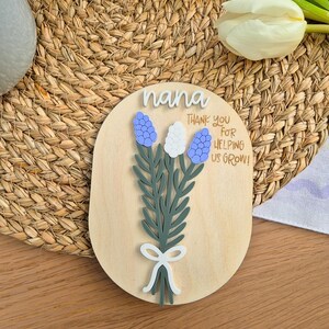 May include: A wooden plaque with the word "nana" in white script. The plaque features a bouquet of blue and white flowers with green leaves and a white bow. The text "THANK YOU FOR HELPING US GROW!" is also on the plaque.