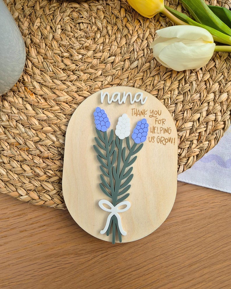 May include: A wooden plaque with the word "nana" in white script. The plaque features a bouquet of blue and white flowers with green leaves and a white bow. The text "THANK YOU FOR HELPING US GROW!" is also on the plaque.