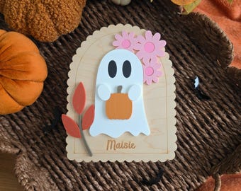 May include: A scalloped wooden plaque featuring a white ghost holding an orange pumpkin, adorned with two pink flowers and a red leaf sprig. The name Maisie is engraved at the bottom of the light-colored wood surface, creating a decorative piece.