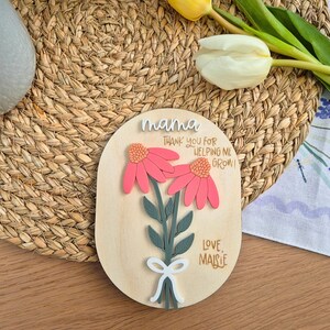May include: Wooden plaque with a floral design. The plaque features two pink flowers with orange centers, green leaves, and a white bow. Text includes "mama", "Thank you for helping me grow!", and "Love, Maisie".