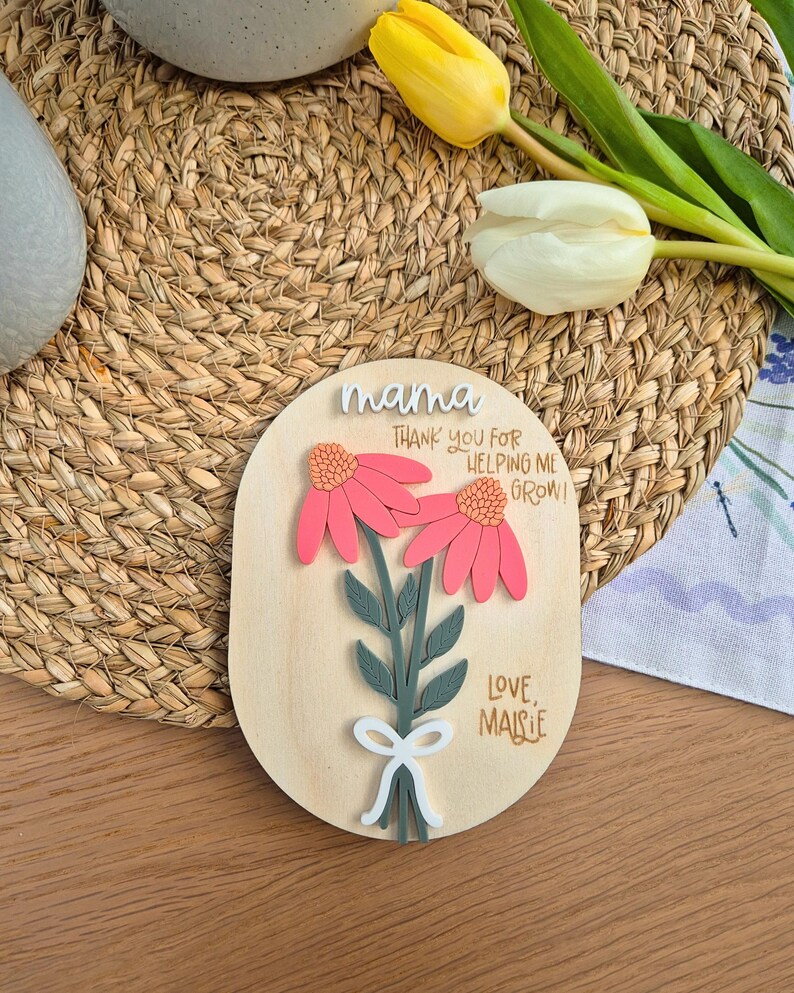 May include: Wooden plaque with a floral design. The plaque features two pink flowers with orange centers, green leaves, and a white bow. Text includes "mama", "Thank you for helping me grow!", and "Love, Maisie".