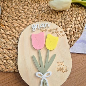 May include: Wooden plaque with a pink and yellow tulip design. The plaque has the word "gran" at the top, with the text "THANK YOU FOR HELPING ME GROW!" and "LOVE, MAISIE" below the flowers. The plaque is on a woven background.