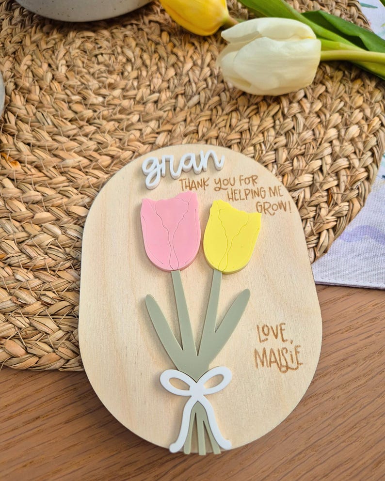 May include: Wooden plaque with a pink and yellow tulip design. The plaque has the word "gran" at the top, with the text "THANK YOU FOR HELPING ME GROW!" and "LOVE, MAISIE" below the flowers. The plaque is on a woven background.