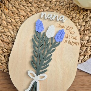 May include: Wooden plaque with a floral design and the word "nana" in white script. The plaque features a bouquet of blue and white flowers with green leaves and a white bow. Text reads "Thank you for helping us grow!"