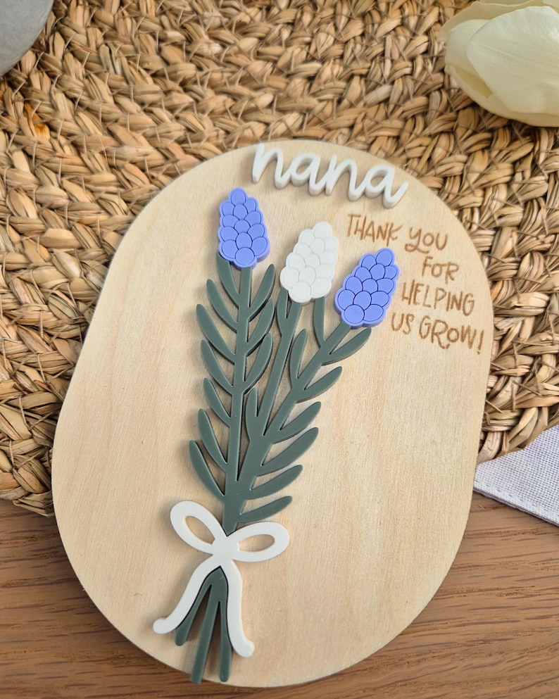 May include: Wooden plaque with a floral design and the word "nana" in white script. The plaque features a bouquet of blue and white flowers with green leaves and a white bow. Text reads "Thank you for helping us grow!"