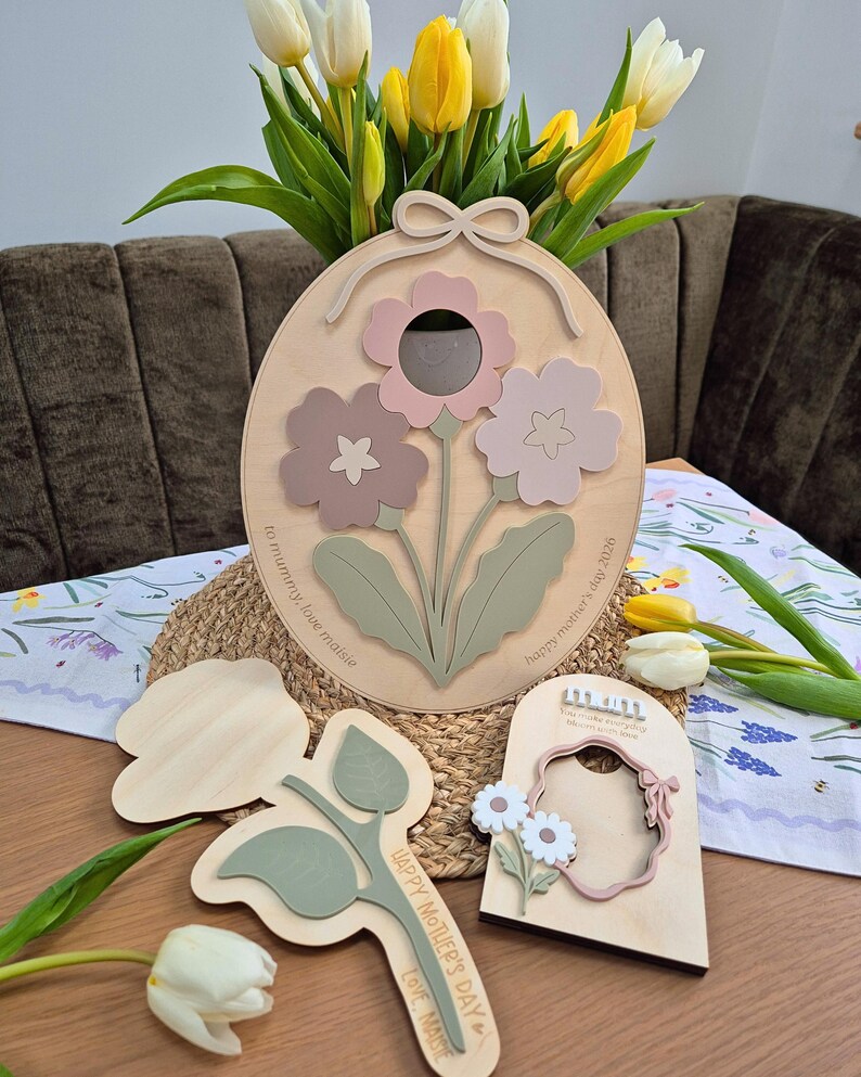 May include: Wooden floral wall art featuring three-dimensional flowers in pink, mauve, and green. The design includes a bow at the top and the text "Happy Mother's Day 2026" and "to mummy love maisie". Additional wooden flower cutouts are also present.