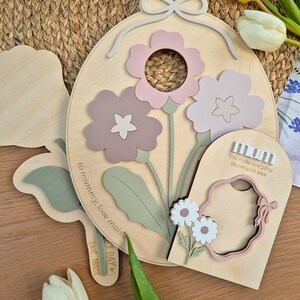 May include: Wooden floral wall art featuring three-dimensional flowers in pink and mauve shades. The design includes a bow, leaves, and the text "You make everyday bloom with love" and "to mummy, love maisie".