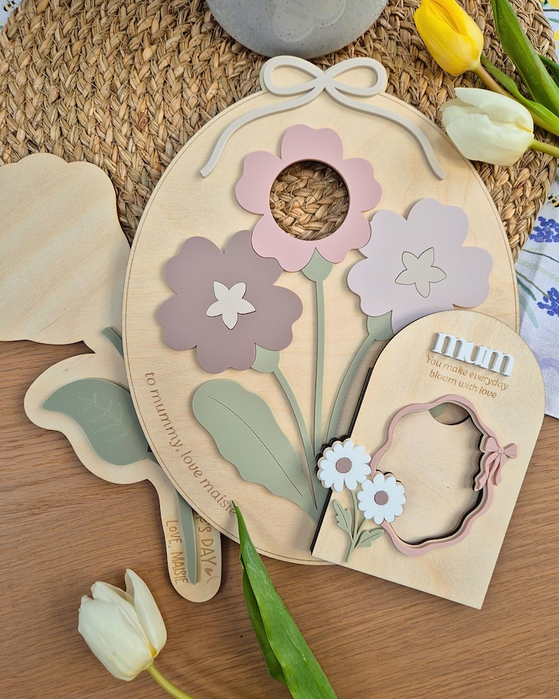 May include: Wooden floral wall art featuring three-dimensional flowers in pink and mauve shades. The design includes a bow, leaves, and the text "You make everyday bloom with love" and "to mummy, love maisie".