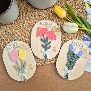 May include: Three wooden floral plaques with the words "mama", "gran", and "nana". Each plaque features a different flower arrangement in pink, yellow, and blue, with the text "Thank you for helping me grow!" and "Love Maisie".