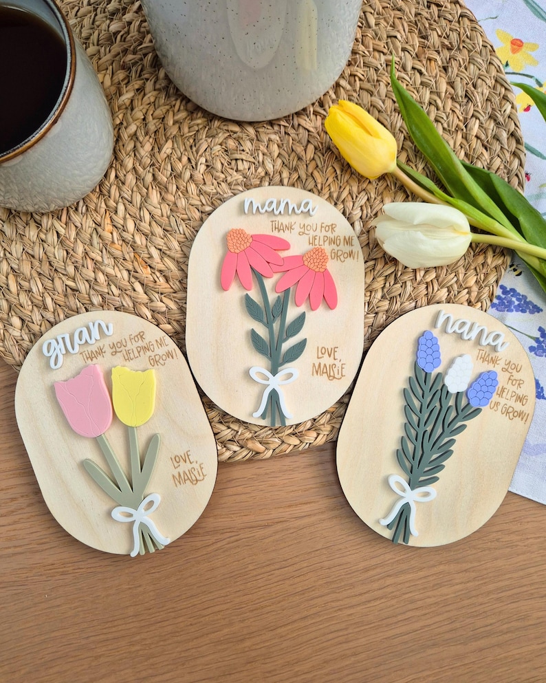 May include: Three wooden floral plaques with the words "mama", "gran", and "nana". Each plaque features a different flower arrangement in pink, yellow, and blue, with the text "Thank you for helping me grow!" and "Love Maisie".