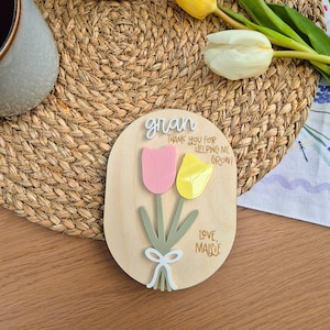 May include: Wooden oval plaque with a pink and yellow tulip design. The plaque has the word "gran" and the message "Thank you for helping me grow! Love, Maisie" in black script. A white bow is at the base of the tulips.
