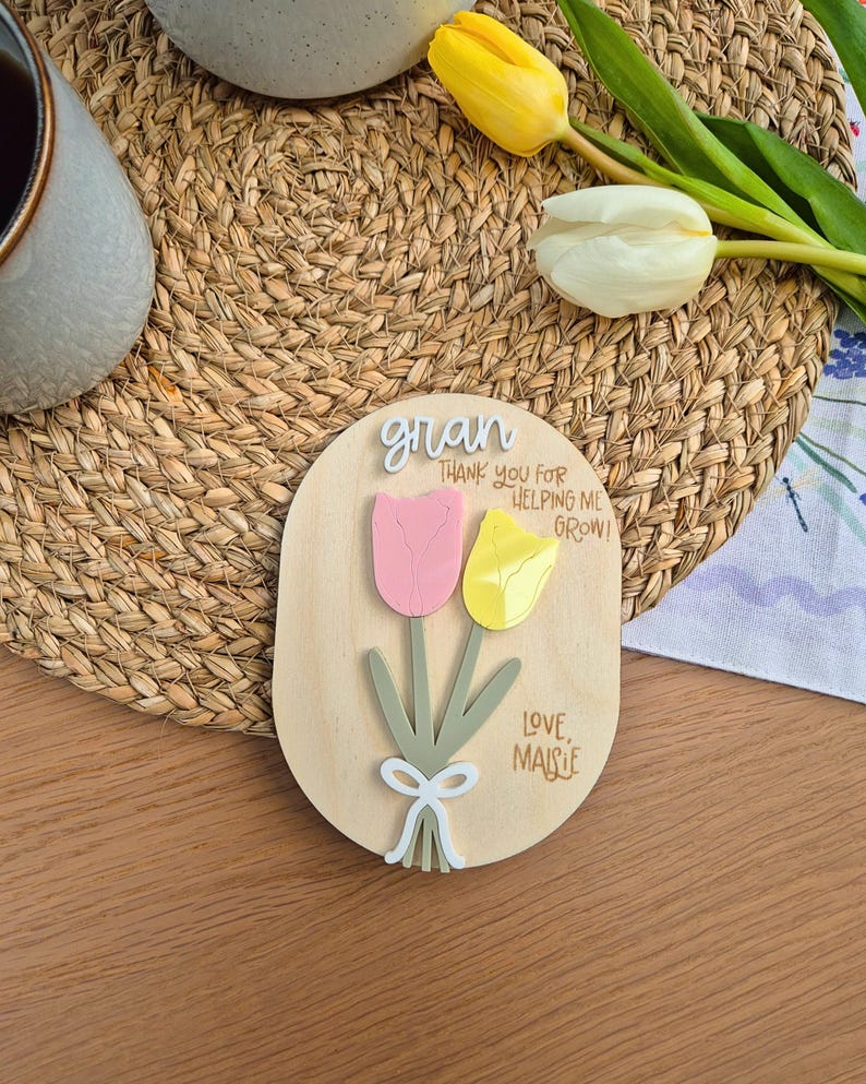 May include: Wooden oval plaque with a pink and yellow tulip design. The plaque has the word "gran" and the message "Thank you for helping me grow! Love, Maisie" in black script. A white bow is at the base of the tulips.