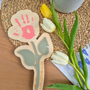 May include: Wooden Mother's Day gift in the shape of a flower, featuring a handprint and the text "Happy Mother's Day, Love, Maisie." A bouquet of white and yellow tulips is next to the gift. The handprint is pink.