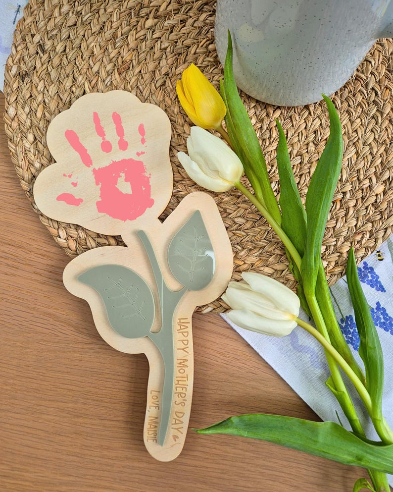 May include: Wooden Mother's Day gift in the shape of a flower, featuring a handprint and the text "Happy Mother's Day, Love, Maisie." A bouquet of white and yellow tulips is next to the gift. The handprint is pink.