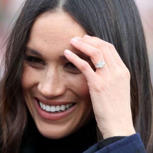 May include: A close-up of a woman smiling, with dark hair and a navy blue coat. A diamond ring with a large center stone and two side stones is on her ring finger. The ring is set in a silver band.