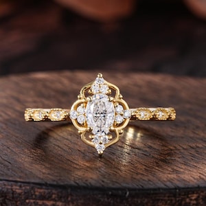 May include: A gold engagement ring with a central oval-cut diamond, surrounded by smaller diamonds in an ornate, filigree setting. The band features delicate detailing. The ring is displayed on a dark wooden surface.