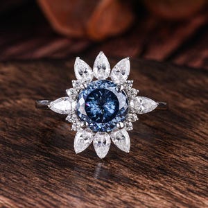 May include: A silver ring featuring a large, round, blue gemstone surrounded by smaller white gemstones in a floral pattern. The ring is set on a dark wooden surface, creating a contrast of colors and textures.
