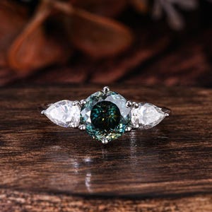 May include: A three-stone ring featuring a round, teal-colored center stone flanked by two pear-shaped, white side stones. The ring is set on a silver band and rests on a dark wooden surface, showcasing the jewelry's design.