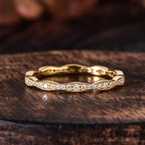 Könnte beinhalten: Ein goldfarbener Ring mit wellenförmigem Design, besetzt mit kleinen, klaren Edelsteinen. Der Ring wird auf einer dunklen Holzoberfläche präsentiert, die seine komplizierten Details und sein Funkeln hervorhebt. Der Ring ist ein Schmuckstück.