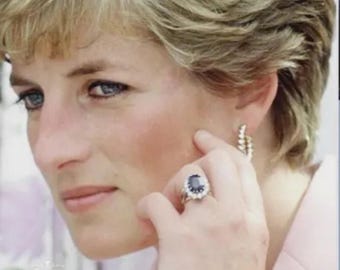 Könnte beinhalten: Nahaufnahme einer Frau, die einen goldenen Ring mit einem großen ovalen blauen Edelstein trägt, der von kleineren weißen Edelsteinen umgeben ist. Sie trägt auch goldene Creolen. Die Frau hat kurzes blondes Haar und trägt einen rosa Blazer.