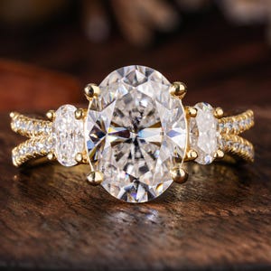 May include: A gold ring with a large, oval-cut clear gemstone as the centerpiece, flanked by two smaller oval stones. The band features a split design with small, sparkling accent stones. The ring is set against a dark wooden surface.