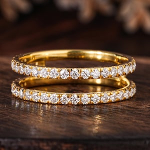 May include: Two gold rings with a continuous row of small, clear gemstones. The rings are set on a dark wooden surface. The gemstones sparkle, reflecting light. The rings are likely for engagement or wedding.