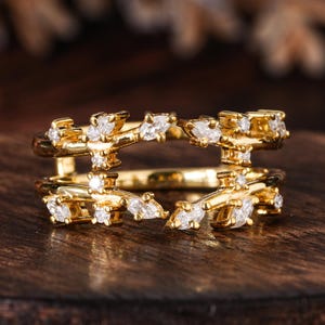 Peut inclure: Une bague dorée avec un design à double bande. La bague présente un motif en forme de branche orné de plusieurs petites pierres précieuses claires. Les pierres précieuses sont serties le long des branches, créant un effet scintillant. La bague est présentée sur une surface en bois.