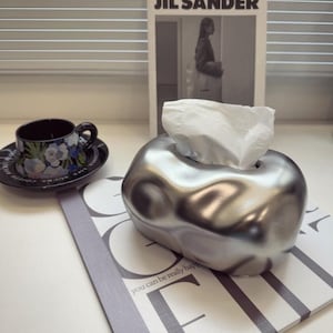 Puede incluir: Un dispensador de pañuelos plateado con una forma orgánica única, que dispensa pañuelos blancos. Una taza negra con diseños florales descansa sobre un platillo. Una revista con el texto "JIL SANDER" está en el fondo.