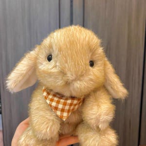 Puede incluir: Un peluche de conejo de color beige claro, con orejas caídas y un pequeño pañuelo a cuadros naranja y blanco. El conejo de peluche tiene ojos negros y pelaje suave. El juguete está diseñado para parecerse a un conejo.