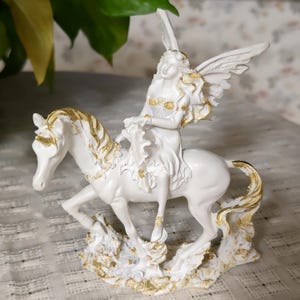 May include: A white figurine of a fairy riding a white horse, with gold accents. The fairy has wings and is wearing a white dress with gold detailing. The horse's mane and tail are also detailed with gold. The base of the figurine is white with gold.