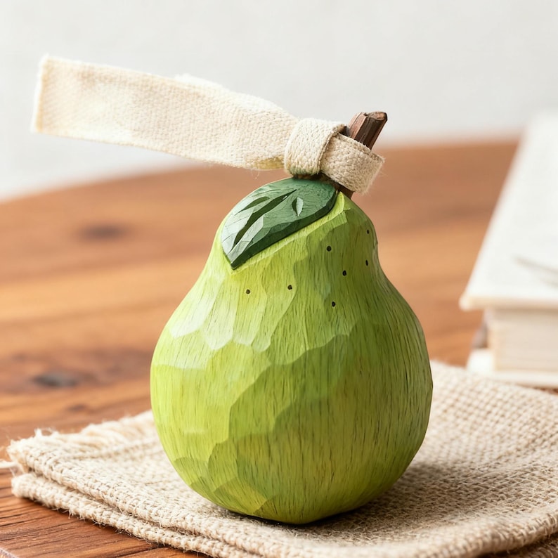 Peut inclure: Figurine de poire en bois sculpt&eacute; &agrave; la main, dans des tons de vert, avec une surface textur&eacute;e et un petit d&eacute;tail de feuille. Un ruban de tissu beige est nou&eacute; autour de la tige. La poire repose sur un tissu de jute.